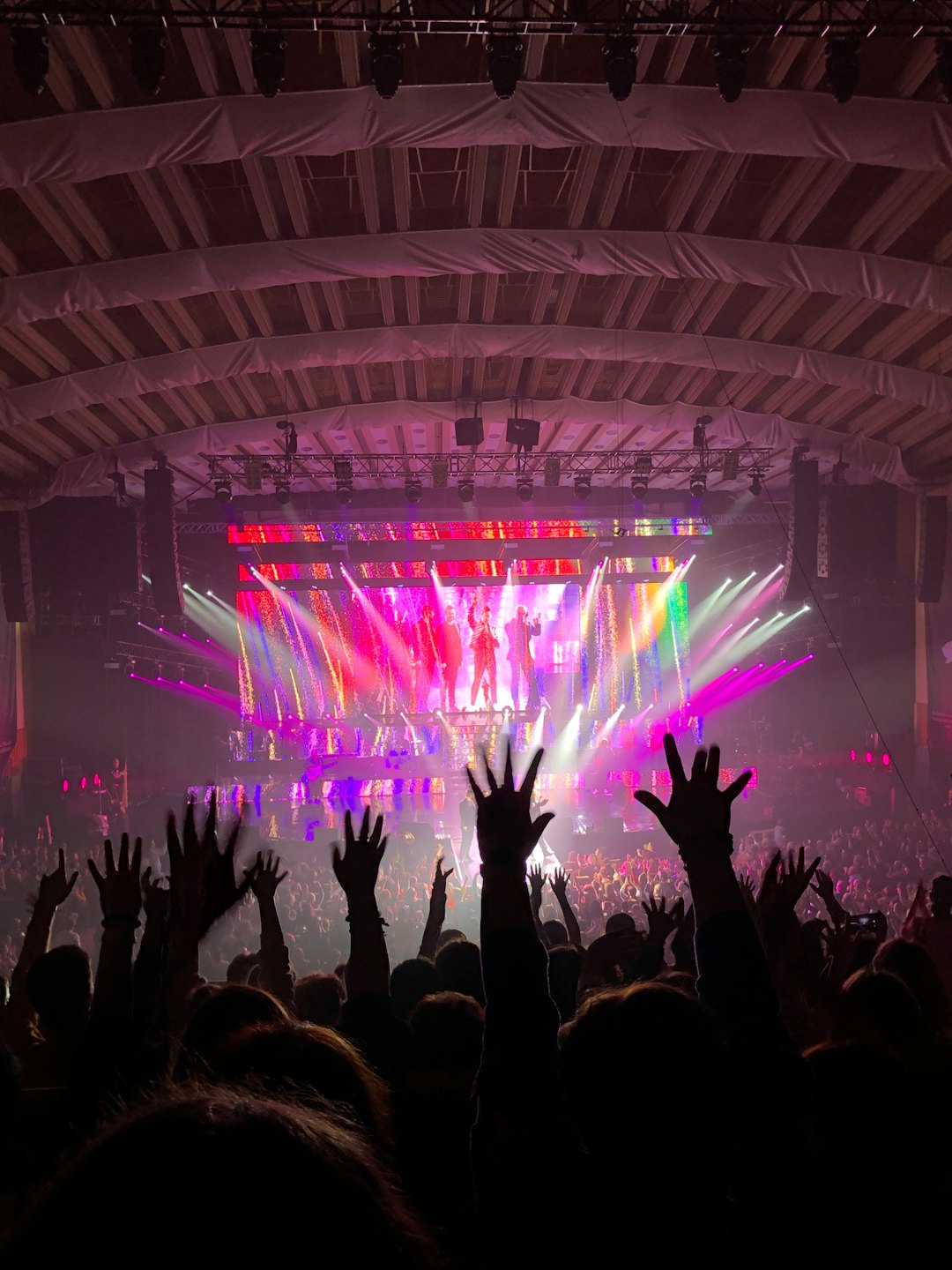 Concert audience with raised hands facing a brightly lit stage.
