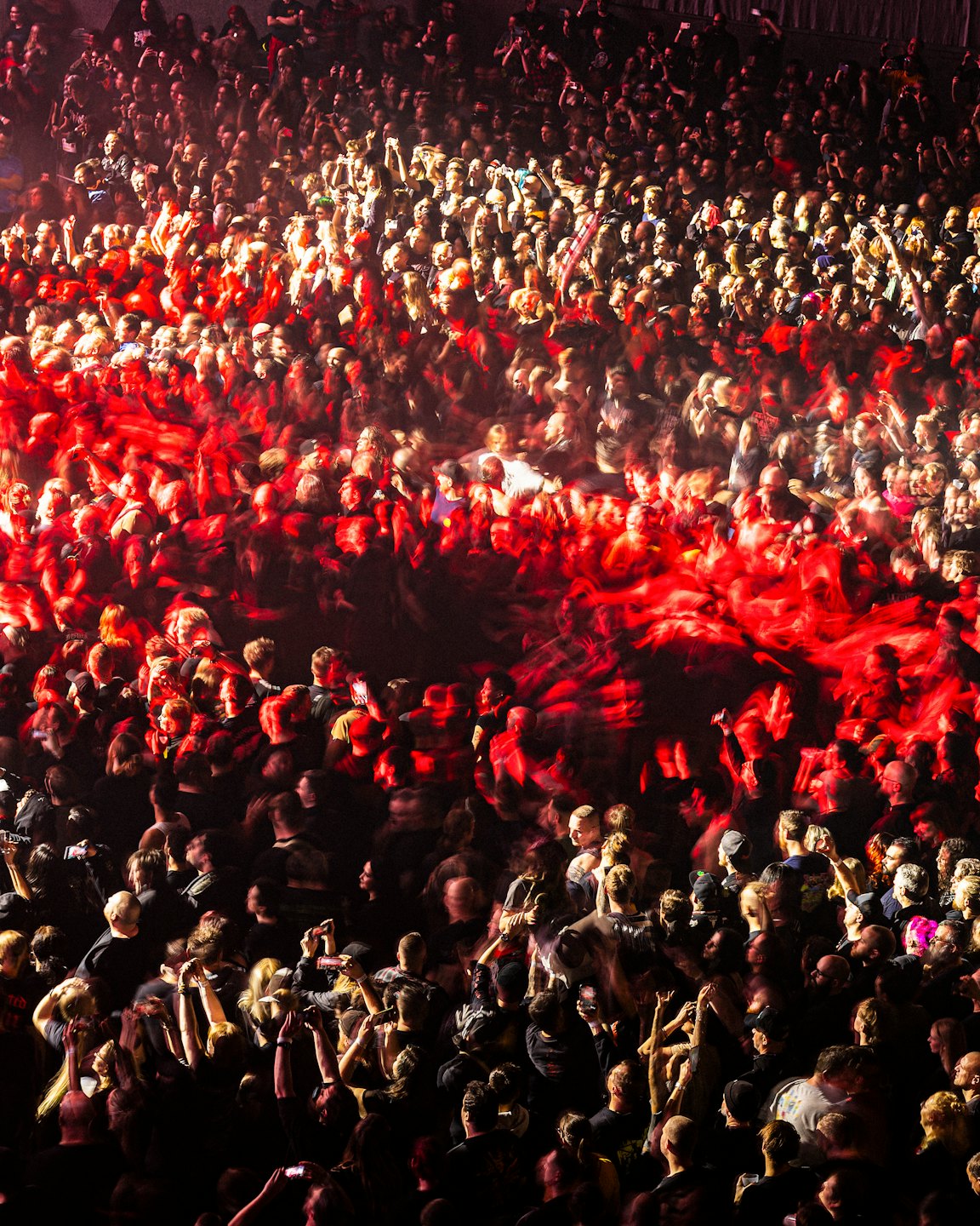 Diverse crowd cheering and recording at a concert under red lights.