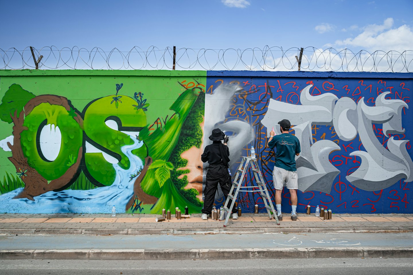 Two artists paint a detailed mural on an outdoor wall.